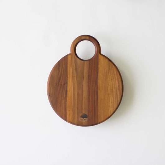 okrugla drvena daska za sečenje i serviranje sa kružnom drškom na vrhu. Napravljena je od ukombinovanih delova različitih boja drveta. Ovako ukombinovani delovi daju dasci prugasti izgled. Uslikana je na beloj podlozi.